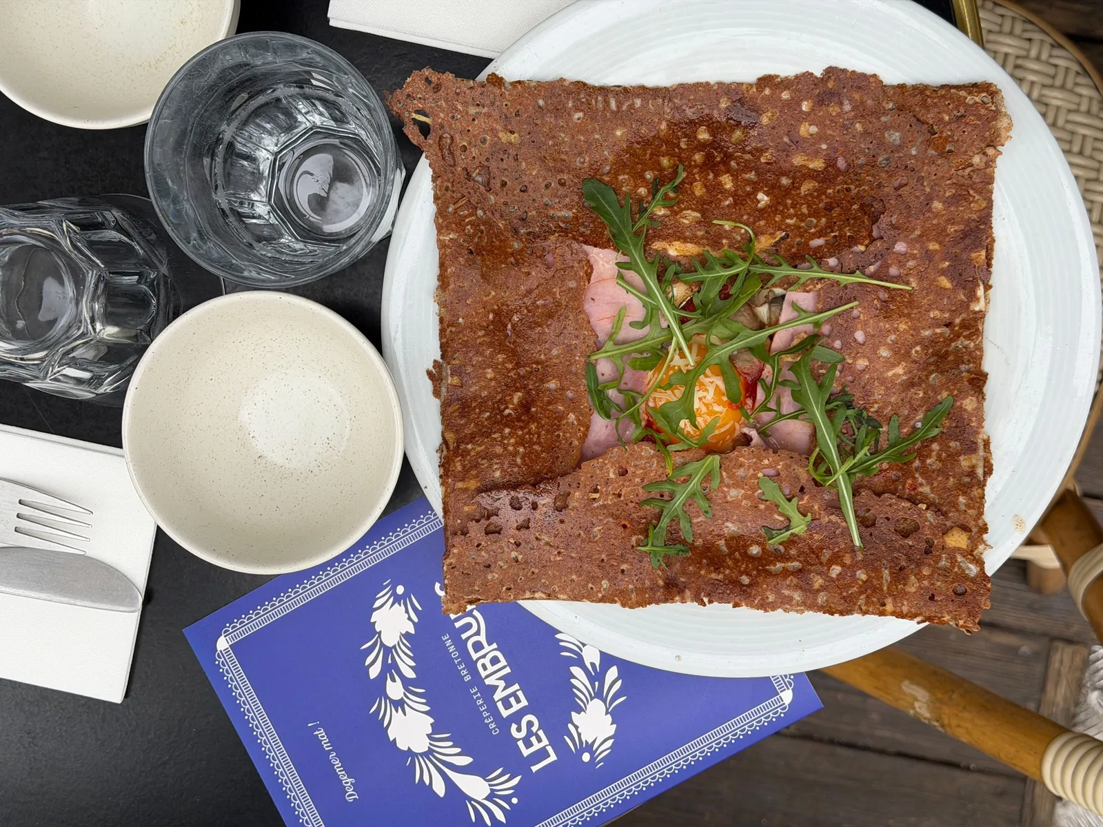 Galette complète au sarrasin, roquette et œuf, posée à côté de la carte bleue Les Embruns